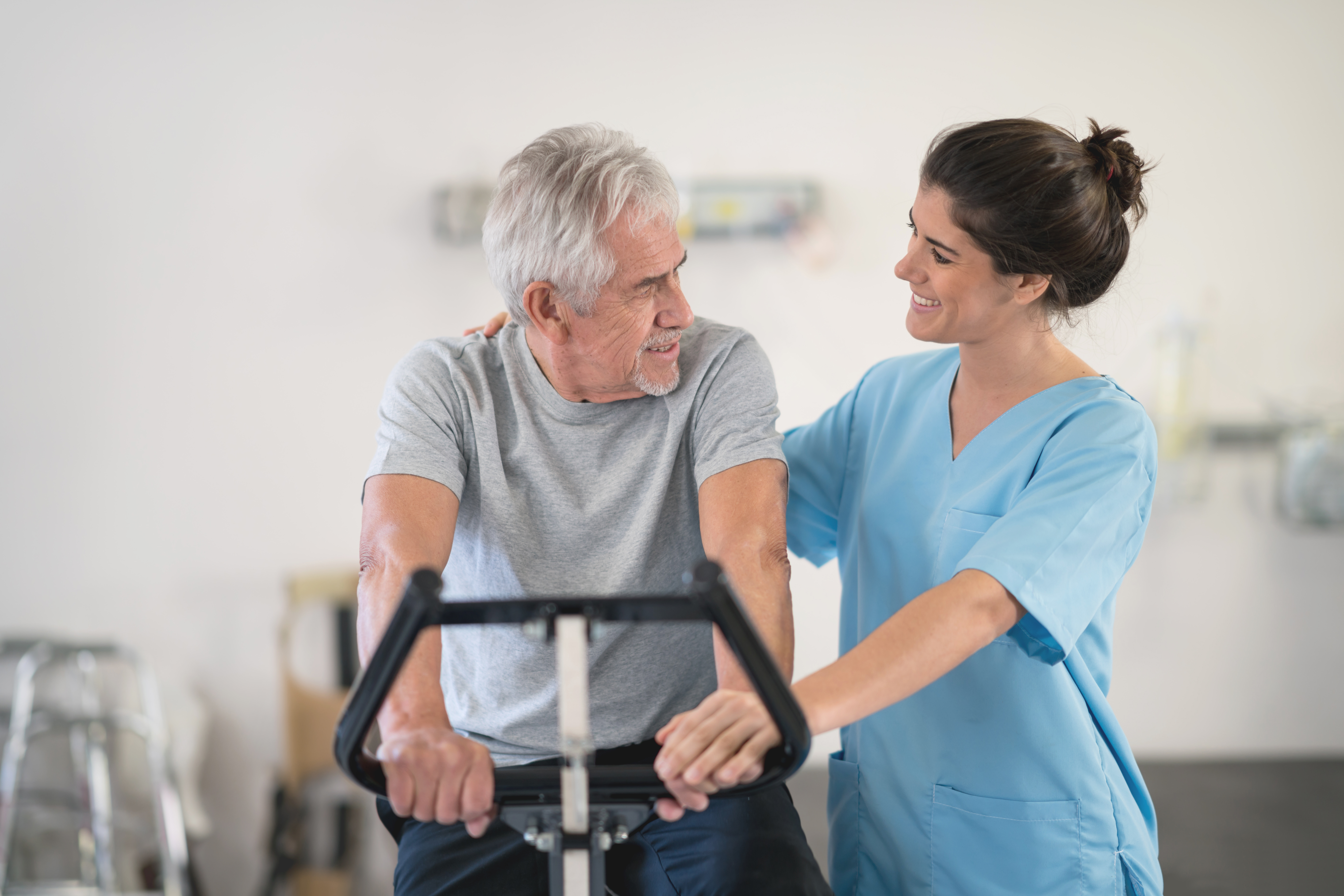 Physiotherapist motivating a senior man on a static bicycle looking at each other smiling. The man had a heart attack and is now at a skilled nursing facility in Bossier City, LA, getting recovery care.